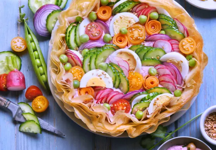 Un plat d'été raffiné : une tarte salée garnie de concombre, radis, œufs durs, tomates cerises, petits pois et oignons rouges, le tout sur un lit de crème rose.