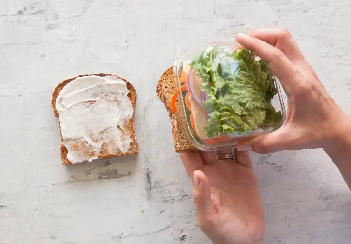 Préparation d'un sandwich crudité : une tranche de pain tartinée de mayonnaise, l'autre garnie de salade, carottes, oignons et fromage, prête à être assemblée.