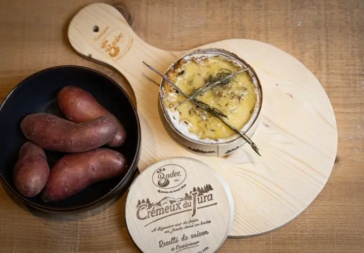 Un crémeux du Jura au four, doré et fondant, avec des pommes de terre rouges dans un bol. Une planche en bois ornée du logo "Crémeux du Jura" complète la scène.