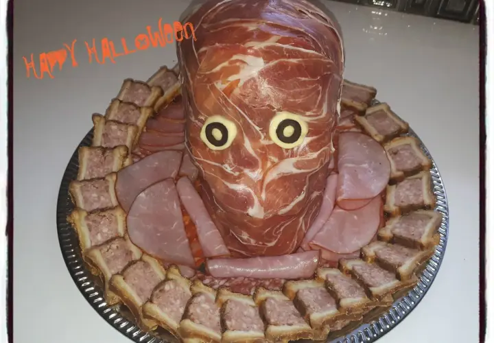 Un plateau de crudités d'Halloween en forme de tête de momie, entouré de petits sandwichs et de charcuterie.