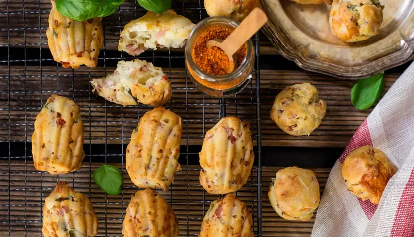 Des madeleines salées originales, garnies de lardons et d'herbes, sur une grille avec un pot d'épices et des feuilles de basilic.