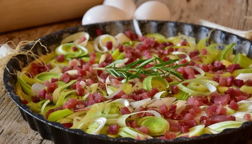 Tarte aux poireaux et lardons, une idée savoureuse pour accompagner des poireaux. Des œufs et un rouleau à pâtisserie sont visibles en arrière-plan.