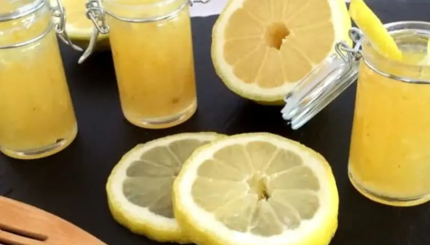 Trois pots de confiture de citron à la mentonnaise, accompagnés de citrons frais coupés et d'une spatule en bois.