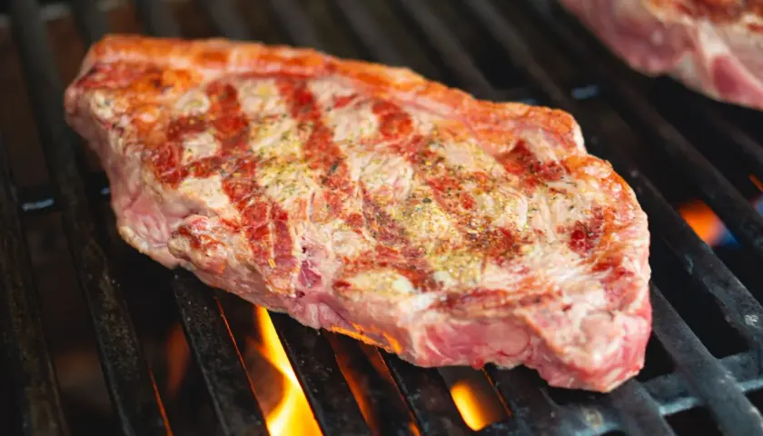 Un steak de basse côte, assaisonné, grésille sur un barbecue. Les flammes dansent sous la grille, promettant une cuisson parfaite.