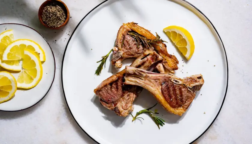Trois côtelettes d'agneau grillées, garnies de romarin frais, avec des quartiers de citron et du poivre concassé en accompagnement.