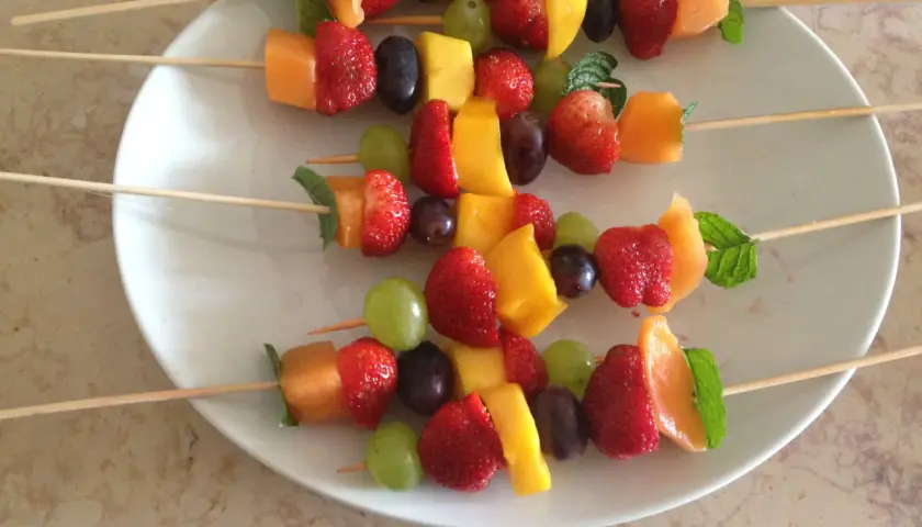 Brochettes de fruits originales : fraises, melon, mangue, raisins et menthe sur des bâtonnets en bois, présentées sur une assiette blanche.