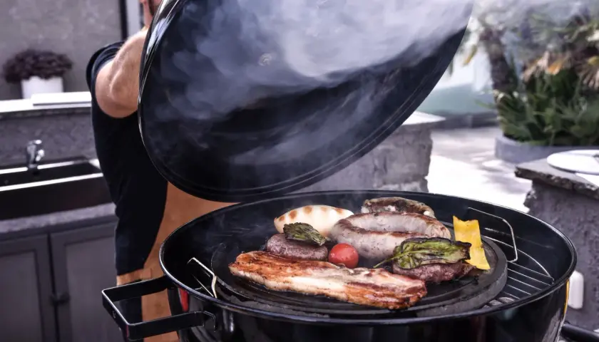 Un homme soulève le couvercle d'un barbecue fumant, révélant une sélection alléchante de quelle viande au tourne broche : saucisses, steaks, lard et légumes grillés.