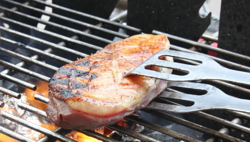 Une aiguillette de canard au barbecue, saisie à la perfection, avec des marques de grill et une peau dorée. Les pinces à barbecue maintiennent la viande sur la grille chaude.