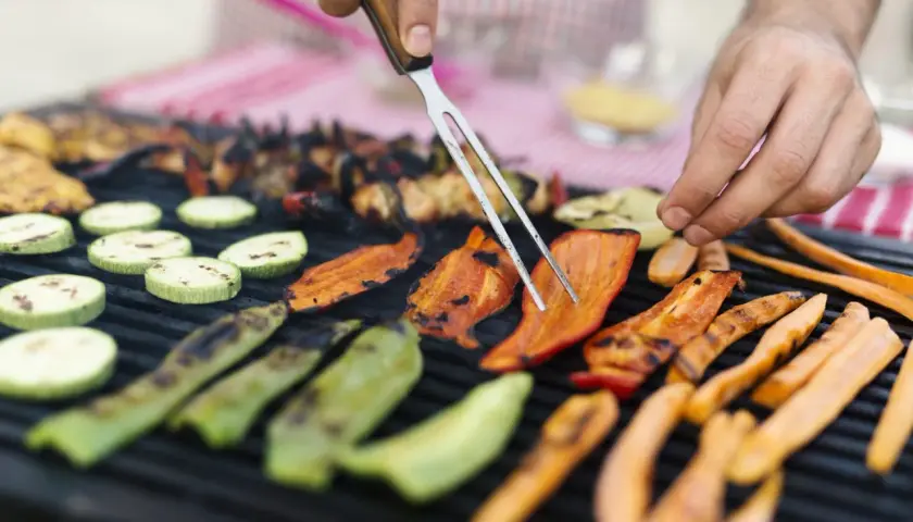 Des légumes accompagnement barbecue grillent sur le feu : courgettes, poivrons et carottes. Un homme les retourne avec une fourchette.
