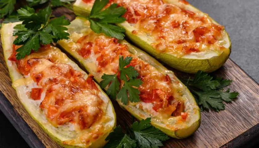 Trois courgettes farcies, façon pizza, gratinées au fromage et garnies de tomates, sur une planche en bois, décorées de persil frais.