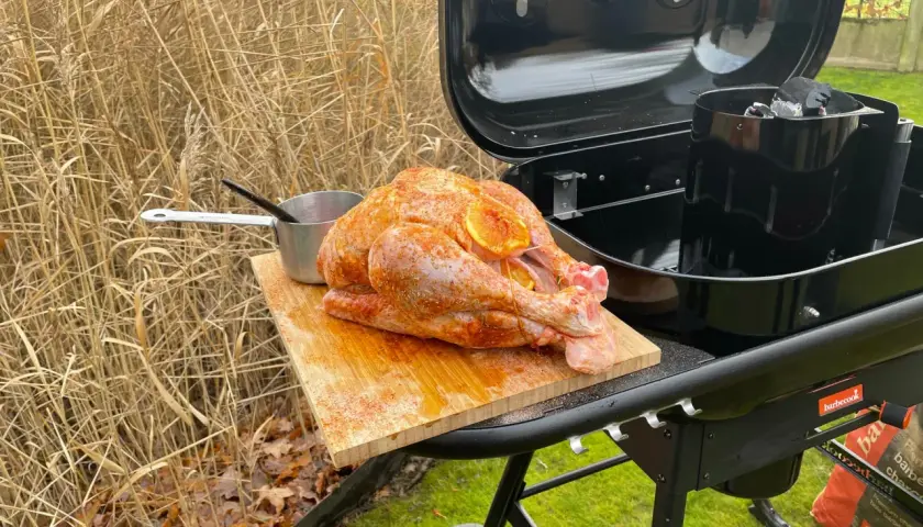 Un dindon assaisonné prêt pour un barbecue de Noël, posé sur une planche à découper à côté d'un barbecue ouvert.