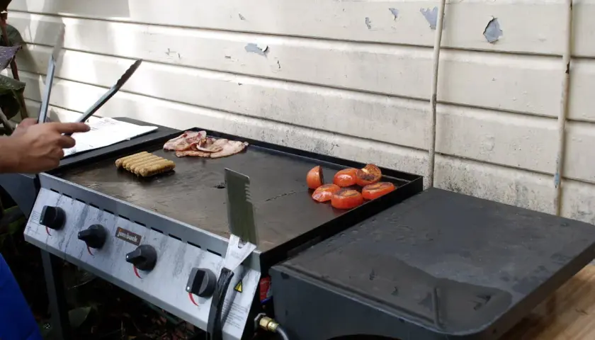 Un barbecue avec une bouteille de gaz prépare un petit-déjeuner savoureux : saucisses, bacon et tomates grillées.