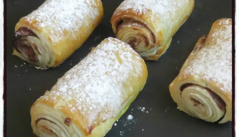Quatre croissants au chocolat en pâte feuilletée, saupoudrés de sucre glace, prêts à être dégustés.