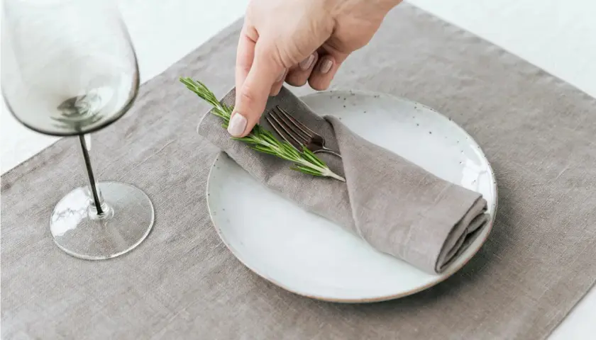Une main dépose une branche de romarin sur un pliage de serviette mariage, sur une assiette blanche.
