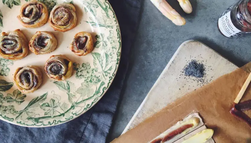 Petits roulés feuilletés à l'asperge blanche, parfaits pour un apéritif. Des asperges blanches, des graines de pavot et une touche de anchois complètent la scène.