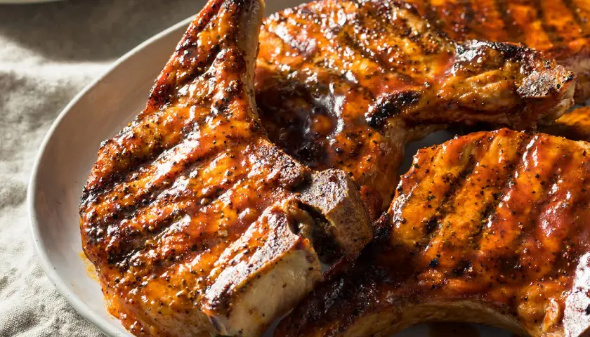 Trois côtes de porcelet grillées, nappées d'une sauce caramélisée et parsemées de poivre noir, sur une assiette blanche.