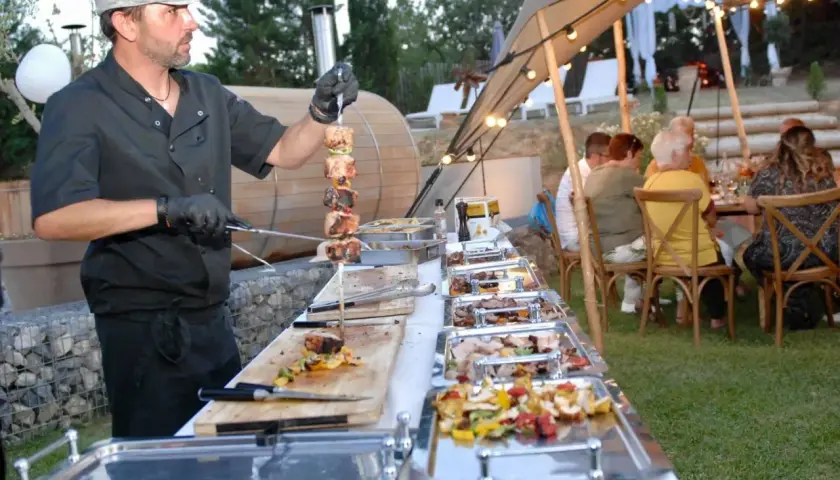 Un chef prépare un menu barbecue mariage. Des brochettes de viande et des légumes sont présentés dans des plats chauffants, tandis que des invités dînent sous une tente décorée de guirlandes lumineuses.