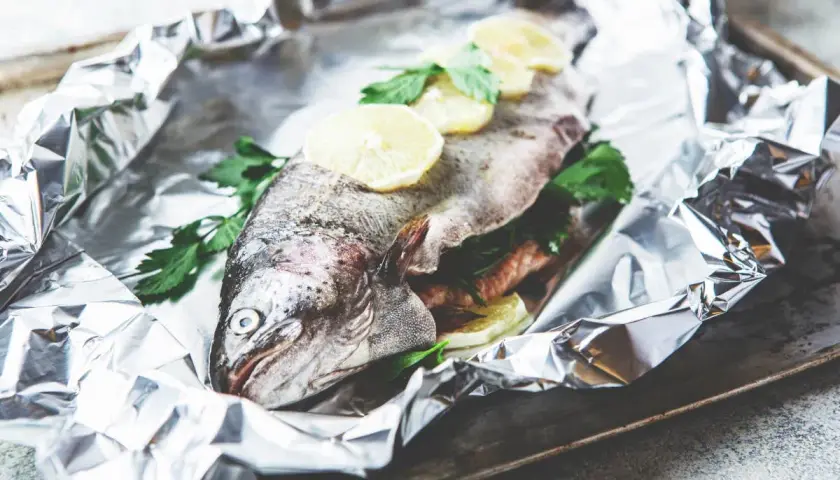 Une truite entière, prête à être cuite en papillote, garnie de rondelles de citron et de persil frais, sur une feuille d'aluminium froissée.