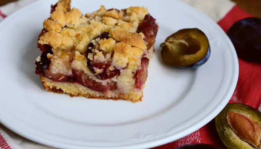 Tranche de gâteau aux prunes à la poêle, garnie de crumble doré et de fruits juteux. Une demi-prune à côté.