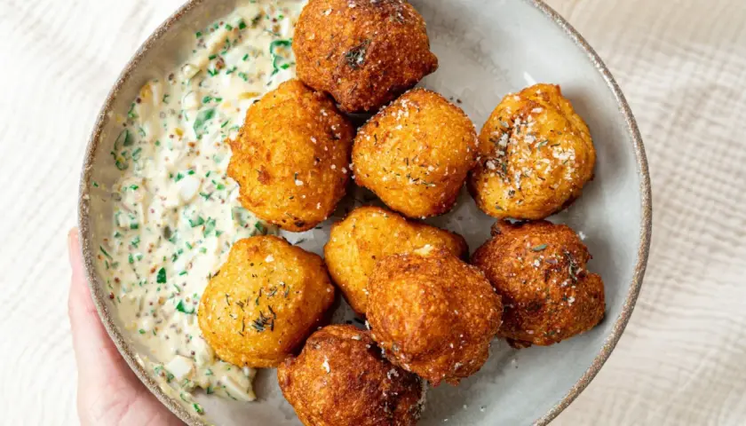 Un bol de pommes dauphines croustillantes, dorées et saupoudrées d'herbes, accompagnées d'une sauce crémeuse aux herbes et aux œufs.