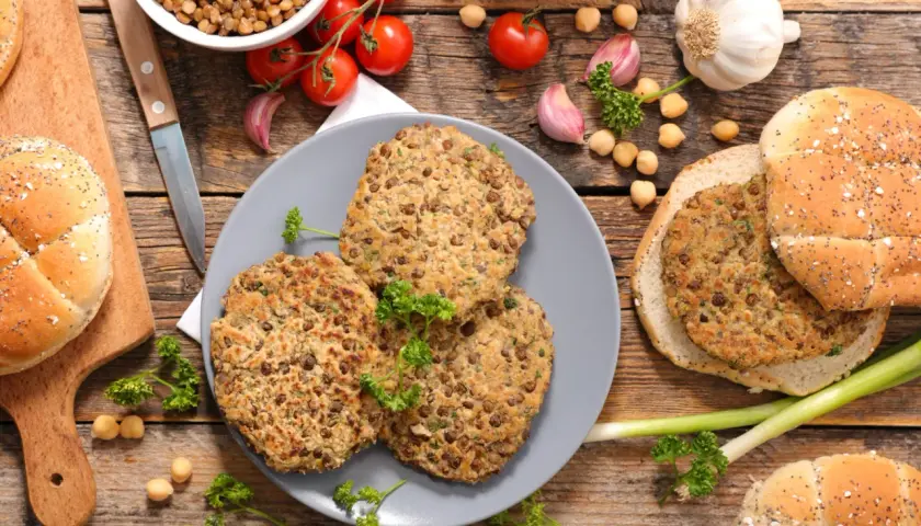 Trois galettes de lentilles, parfaites pour une recette de steak végétarien, sont présentées avec des pains à burger, des tomates et de l'ail sur fond de bois.