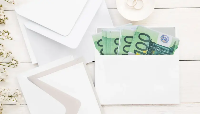 Enveloppes blanches, fleurs et billets de 100€ pour savoir comment offrir de l'argent pour un mariage. Bague de fiançailles sur une soucoupe.