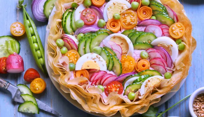 Un plat d'été raffiné : une tarte salée garnie de concombre, radis, œufs durs, tomates cerises, petits pois et oignons rouges, le tout sur un lit de crème rose.