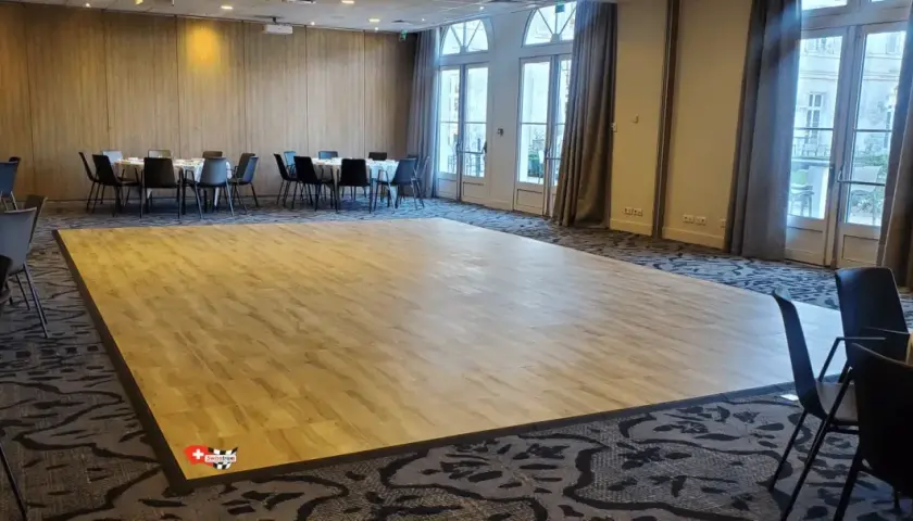 Une piste de danse en bois clair, idéale pour savoir comment faire une piste de danse pas cher. Des tables rondes et des chaises noires sont disposées autour.