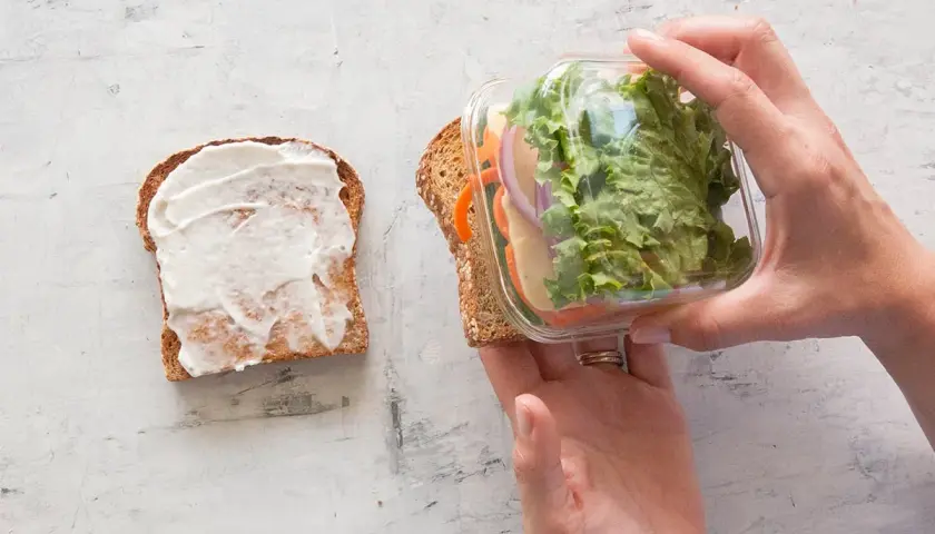 Préparation d'un sandwich crudité : une tranche de pain tartinée de mayonnaise, l'autre garnie de salade, carottes, oignons et fromage, prête à être assemblée.