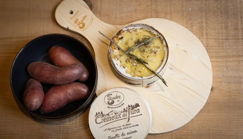 Un crémeux du Jura au four, doré et fondant, avec des pommes de terre rouges dans un bol. Une planche en bois ornée du logo "Crémeux du Jura" complète la scène.