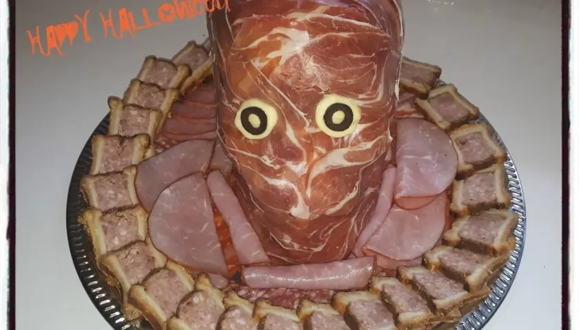 Un plateau de crudités d'Halloween en forme de tête de momie, entouré de petits sandwichs et de charcuterie.