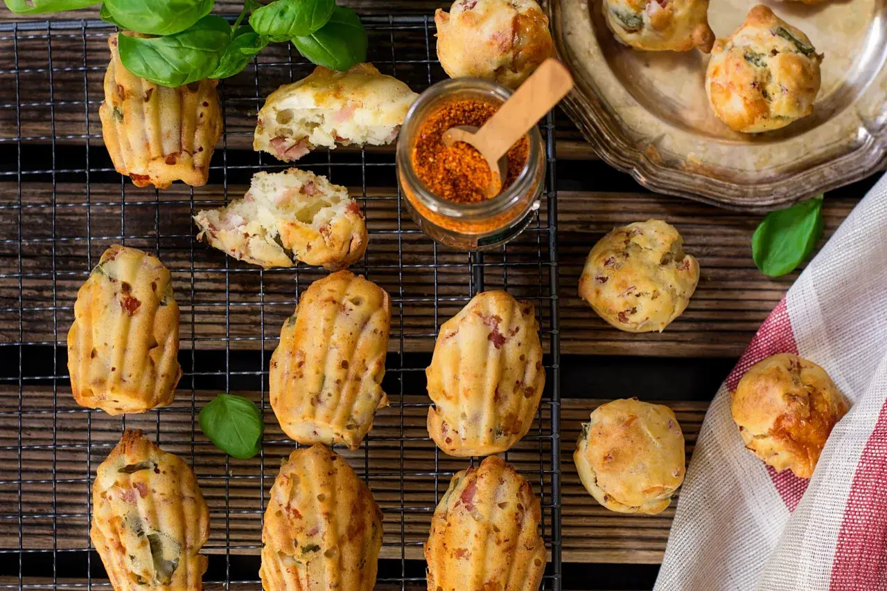 Des madeleines salées originales, garnies de lardons et d'herbes, sur une grille avec un pot d'épices et des feuilles de basilic.