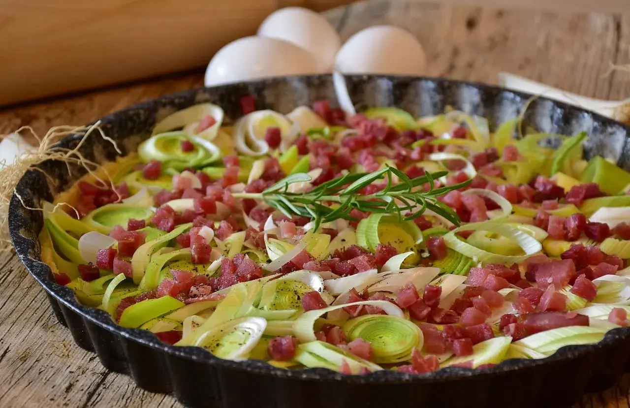 Tarte aux poireaux et lardons, une idée savoureuse pour accompagner des poireaux. Des œufs et un rouleau à pâtisserie sont visibles en arrière-plan.