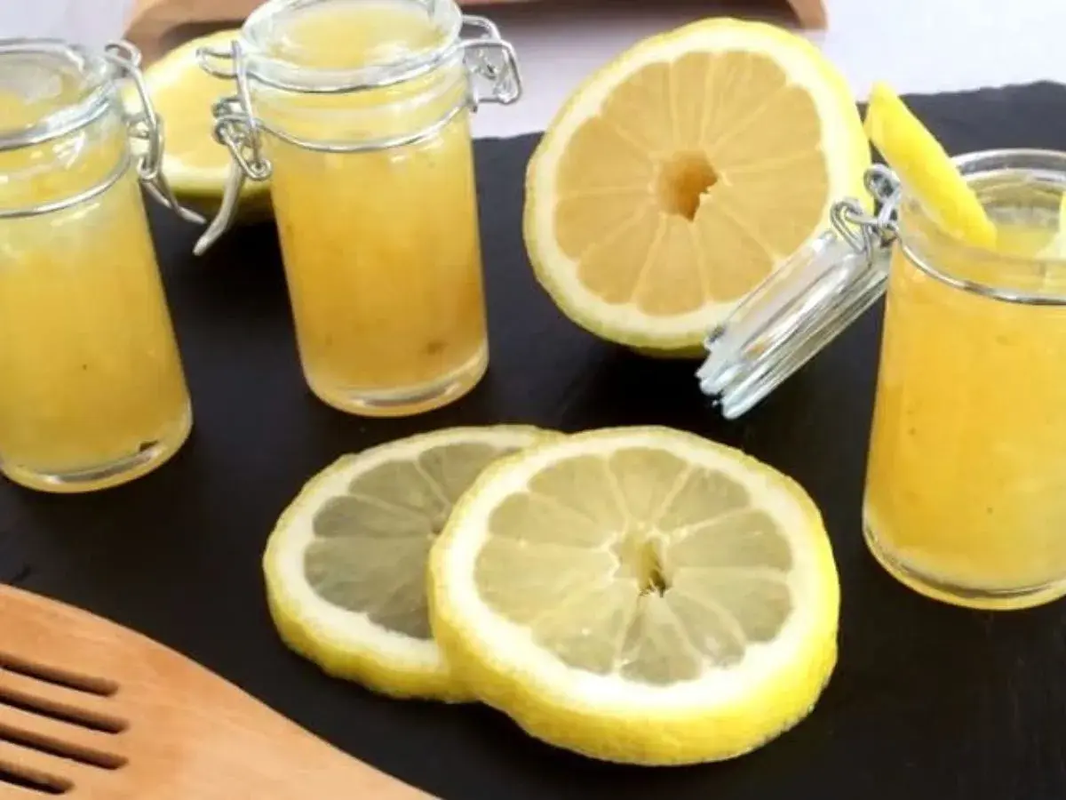 Trois pots de confiture de citron à la mentonnaise, accompagnés de citrons frais coupés et d'une spatule en bois.
