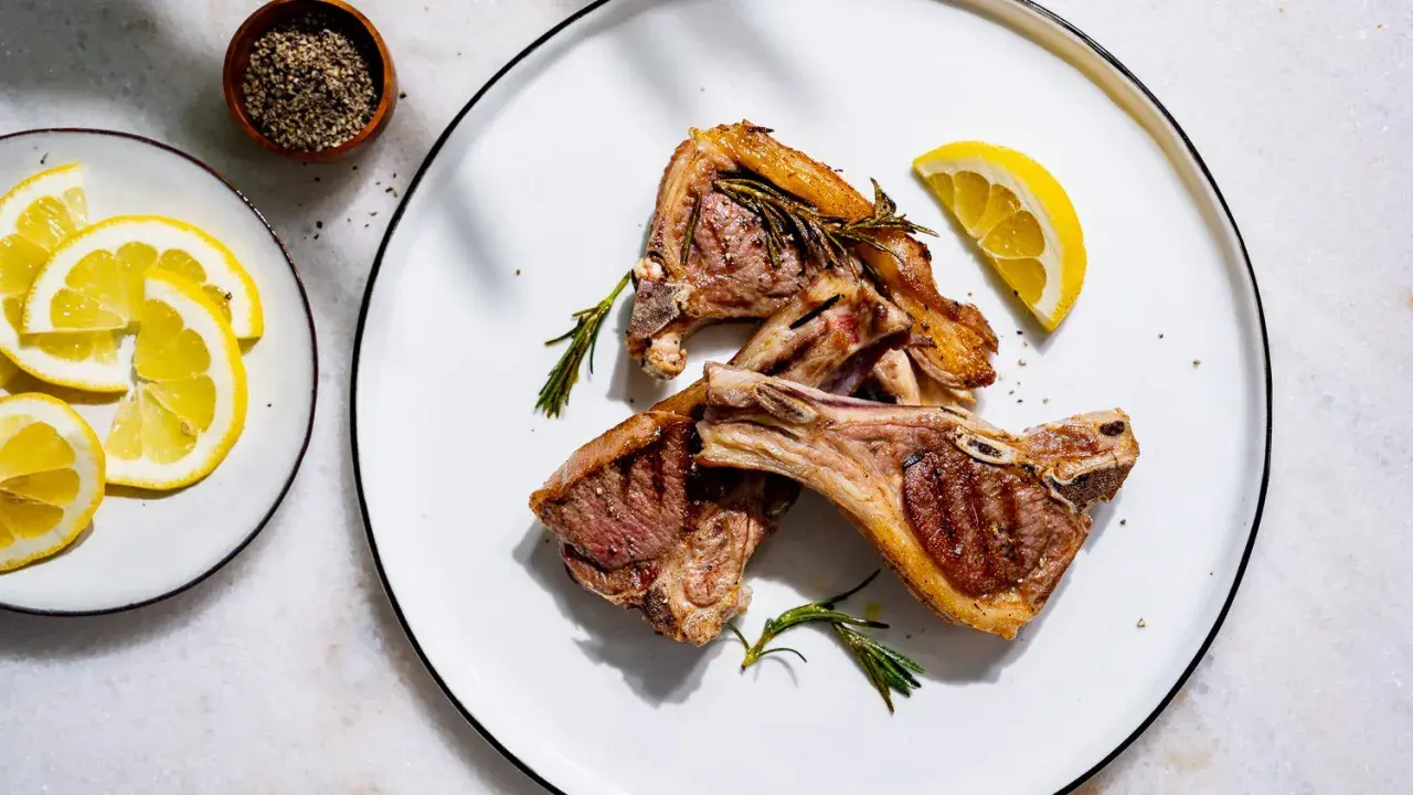 Trois côtelettes d'agneau grillées, garnies de romarin frais, avec des quartiers de citron et du poivre concassé en accompagnement.