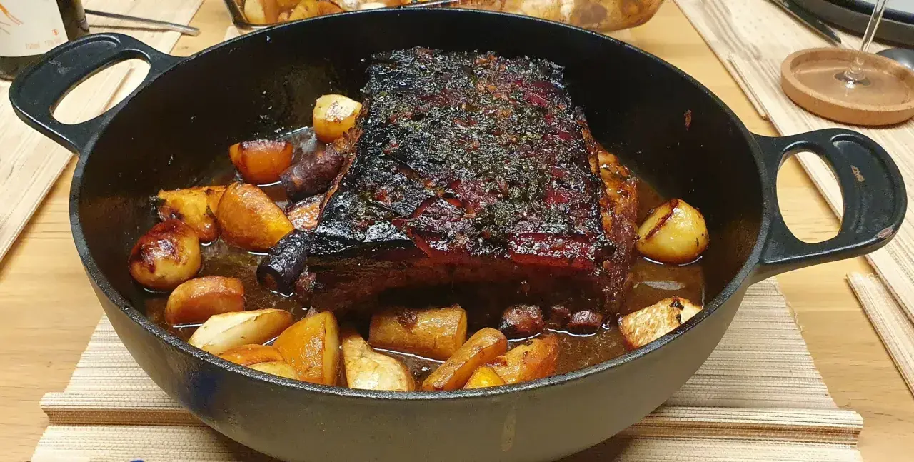Poitrine de porc au four cuisson lente, dorée et savoureuse, accompagnée de pommes de terre rôties dans un plat en fonte.