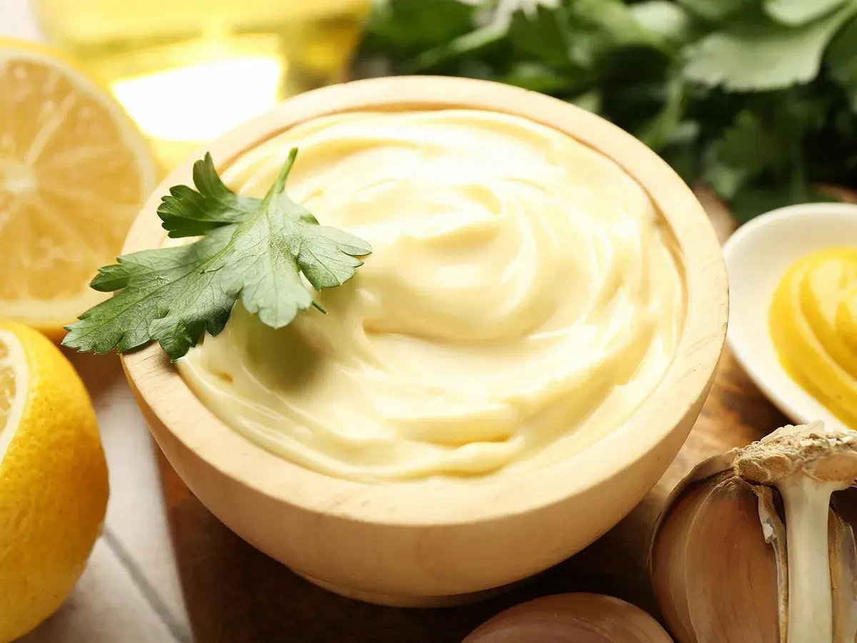 Mayonnaise onctueuse dans un bol en bois, garnie de persil. Citrons, ail et moutarde à proximité. La clé pour ne pas rater une mayonnaise maison.