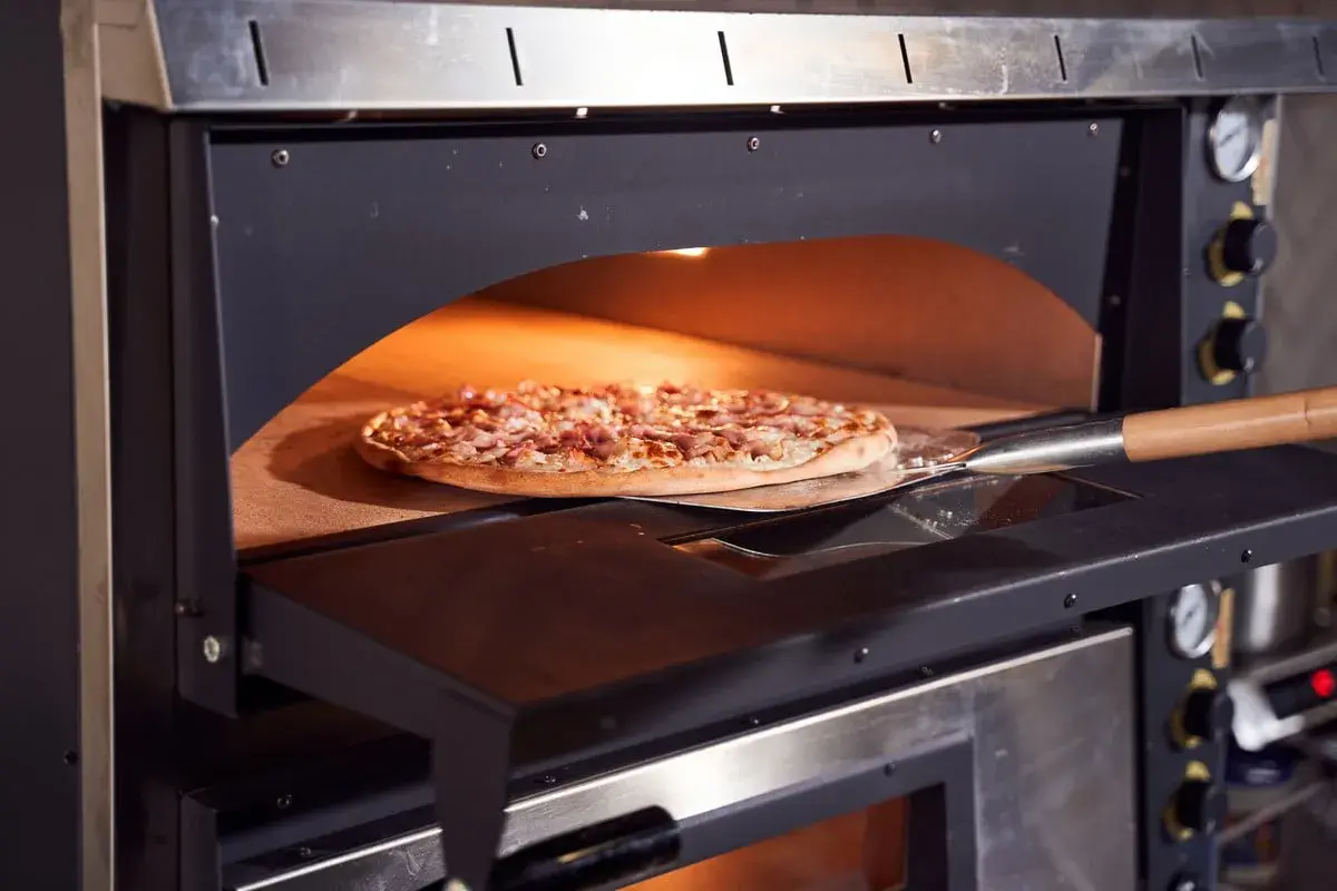 Une pizza garnie est enfournée dans un four à pizza professionnel. La pelle à pizza glisse sous la pâte dorée, prête à être cuite.