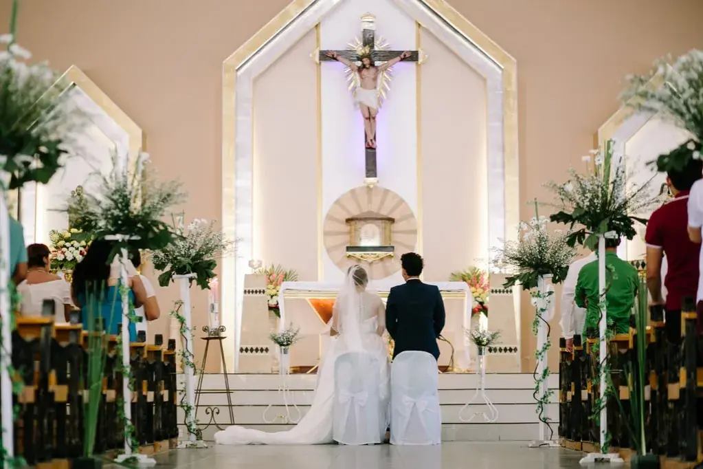Le déroulement du mariage à l'église commence avec le couple devant l'autel, sous un crucifix. Les invités sont assis dans les bancs décorés de fleurs.
