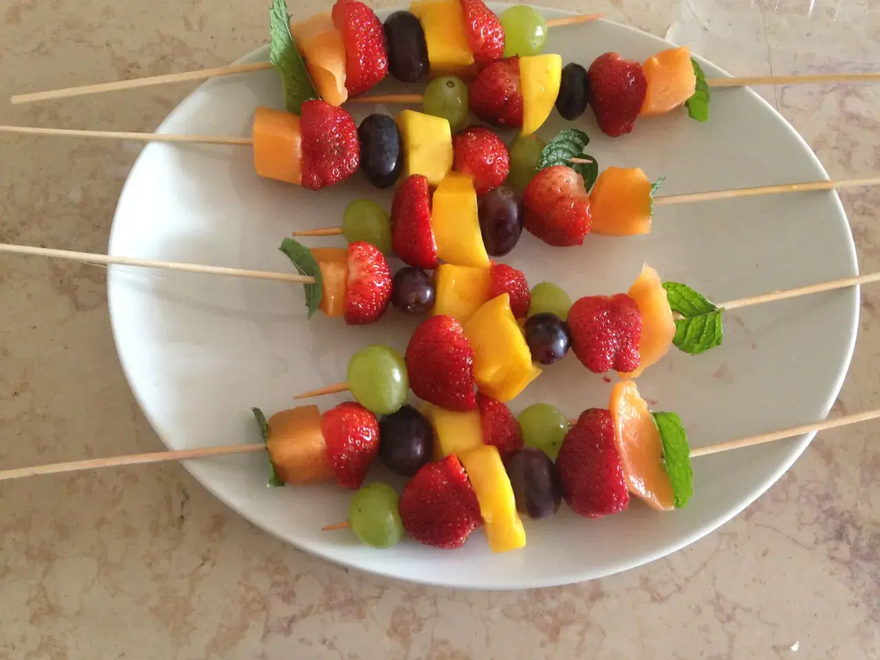 Brochettes de fruits originales : fraises, melon, mangue, raisins et menthe sur des bâtonnets en bois, présentées sur une assiette blanche.
