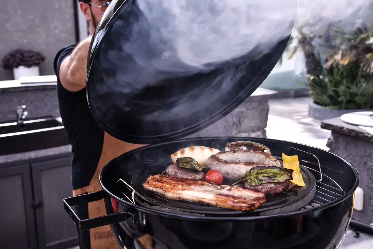 Un homme soulève le couvercle d'un barbecue fumant, révélant une sélection alléchante de quelle viande au tourne broche : saucisses, steaks, lard et légumes grillés.