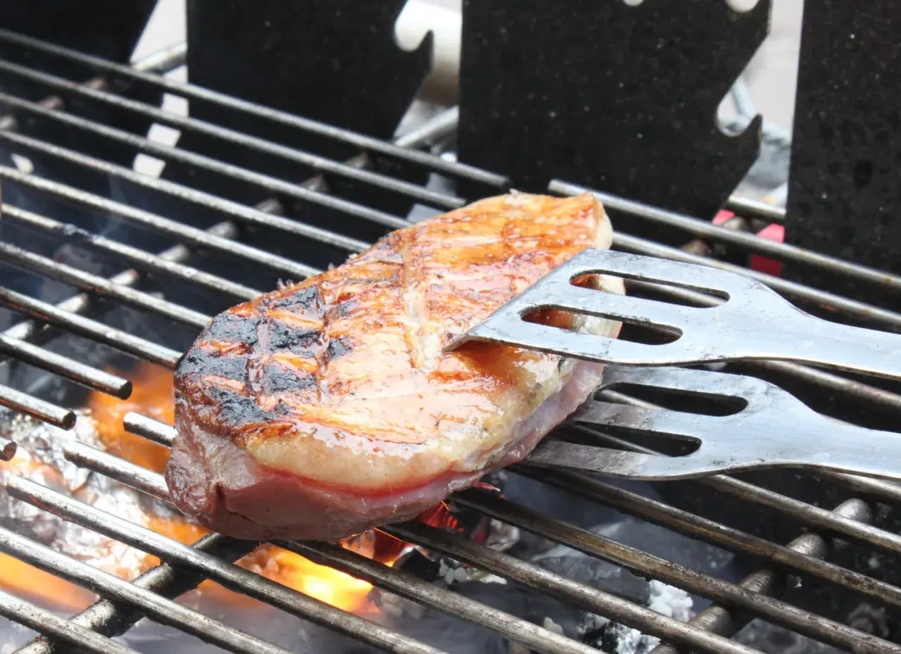Une aiguillette de canard au barbecue, saisie à la perfection, avec des marques de grill et une peau dorée. Les pinces à barbecue maintiennent la viande sur la grille chaude.