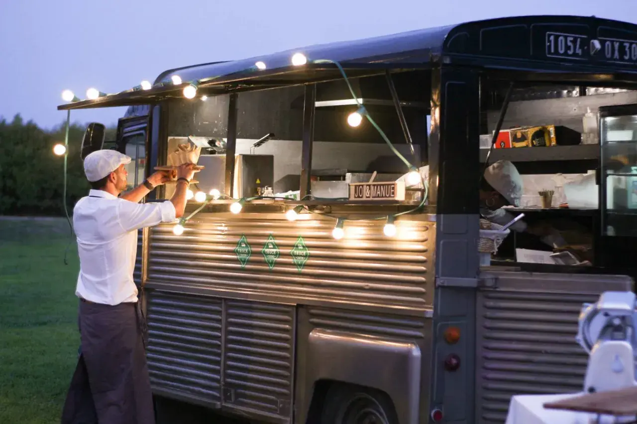 Un food truck illuminé de guirlandes lumineuses, parfait pour une idée food truck mariage. Un serveur sert un plat.