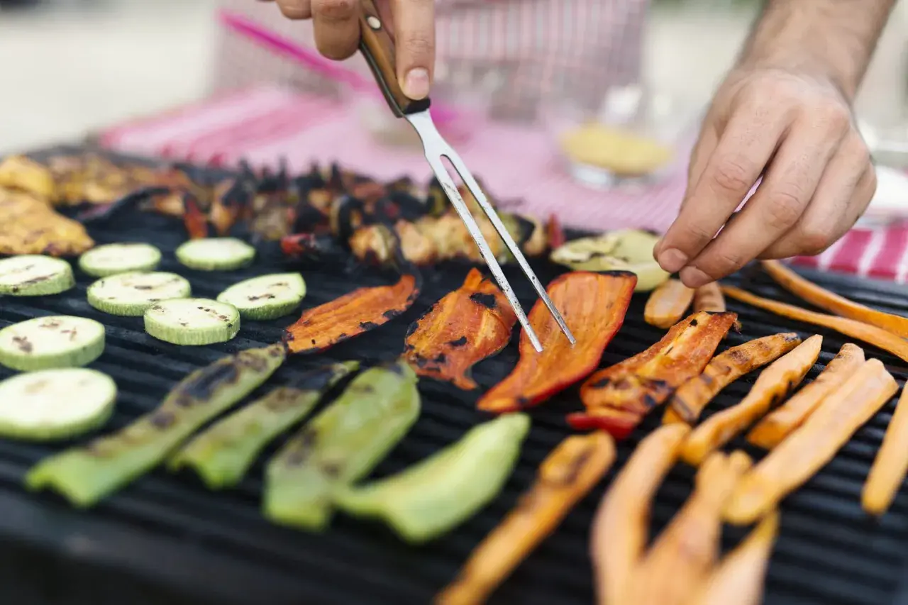 Des légumes accompagnement barbecue grillent sur le feu : courgettes, poivrons et carottes. Un homme les retourne avec une fourchette.