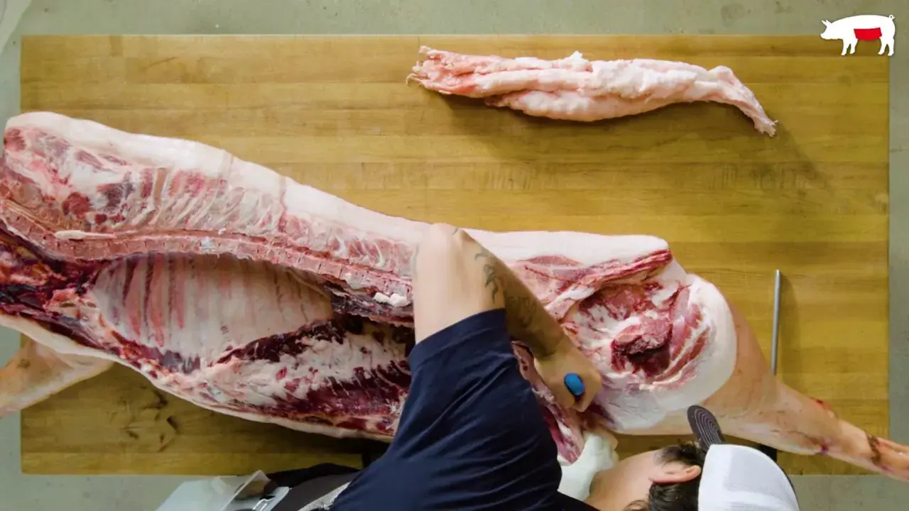 Un boucher démontre comment découper un cochon entier sur une planche en bois. Des morceaux de gras sont déjà séparés.