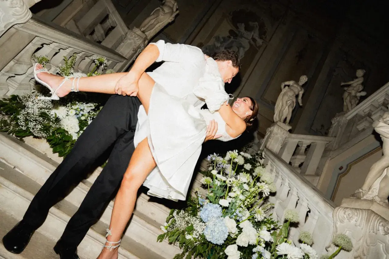 Un couple s'embrasse passionnément lors de leur mariage chez soi, entouré de fleurs blanches et bleues sur un escalier orné de statues.