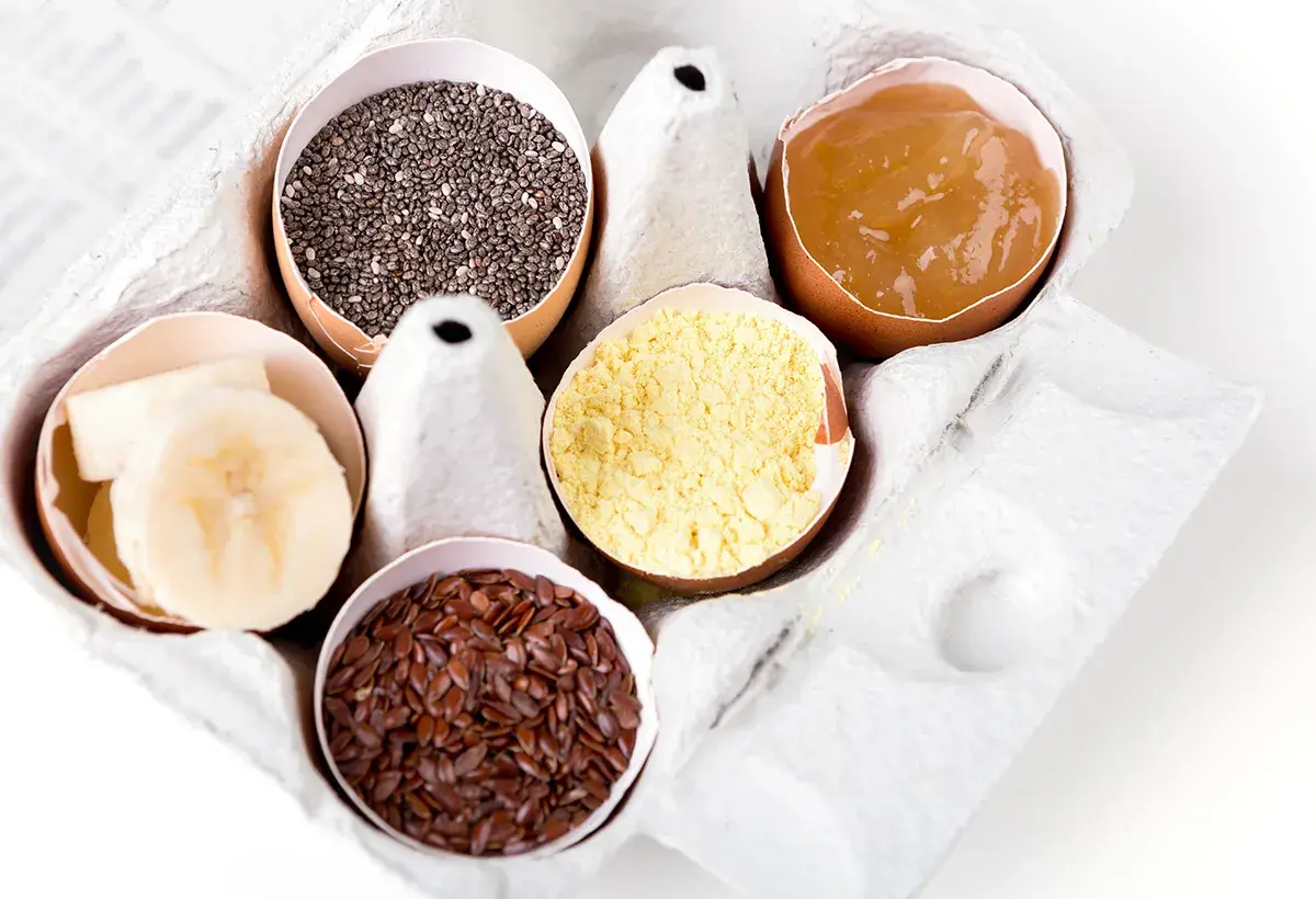 Dans une boîte à œufs, des alternatives pour remplacer les œufs : graines de chia, compote de pommes, farine de pois chiche, graines de lin, rondelles de banane.