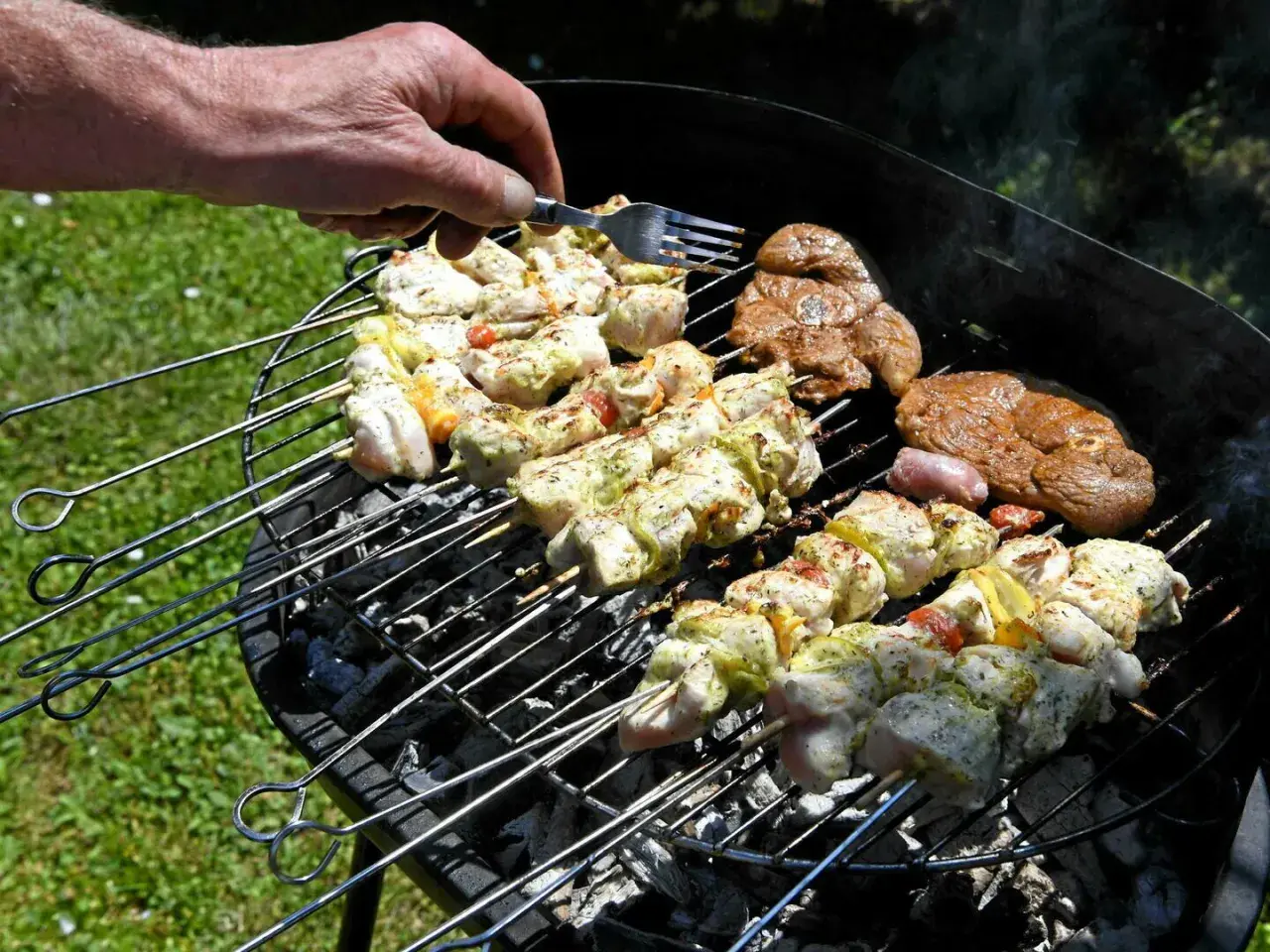 Une main vérifie des brochettes de poulet et des steaks sur un barbecue rustique.