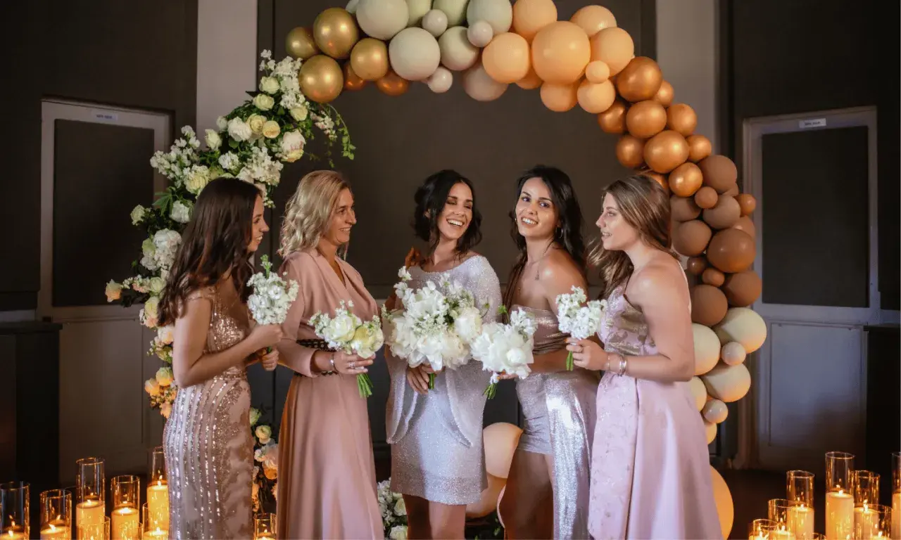 Cinq femmes souriantes, entourées de fleurs et de ballons, se préparent pour un mariage. Elles tiennent des bouquets, prêtes pour la cérémonie.