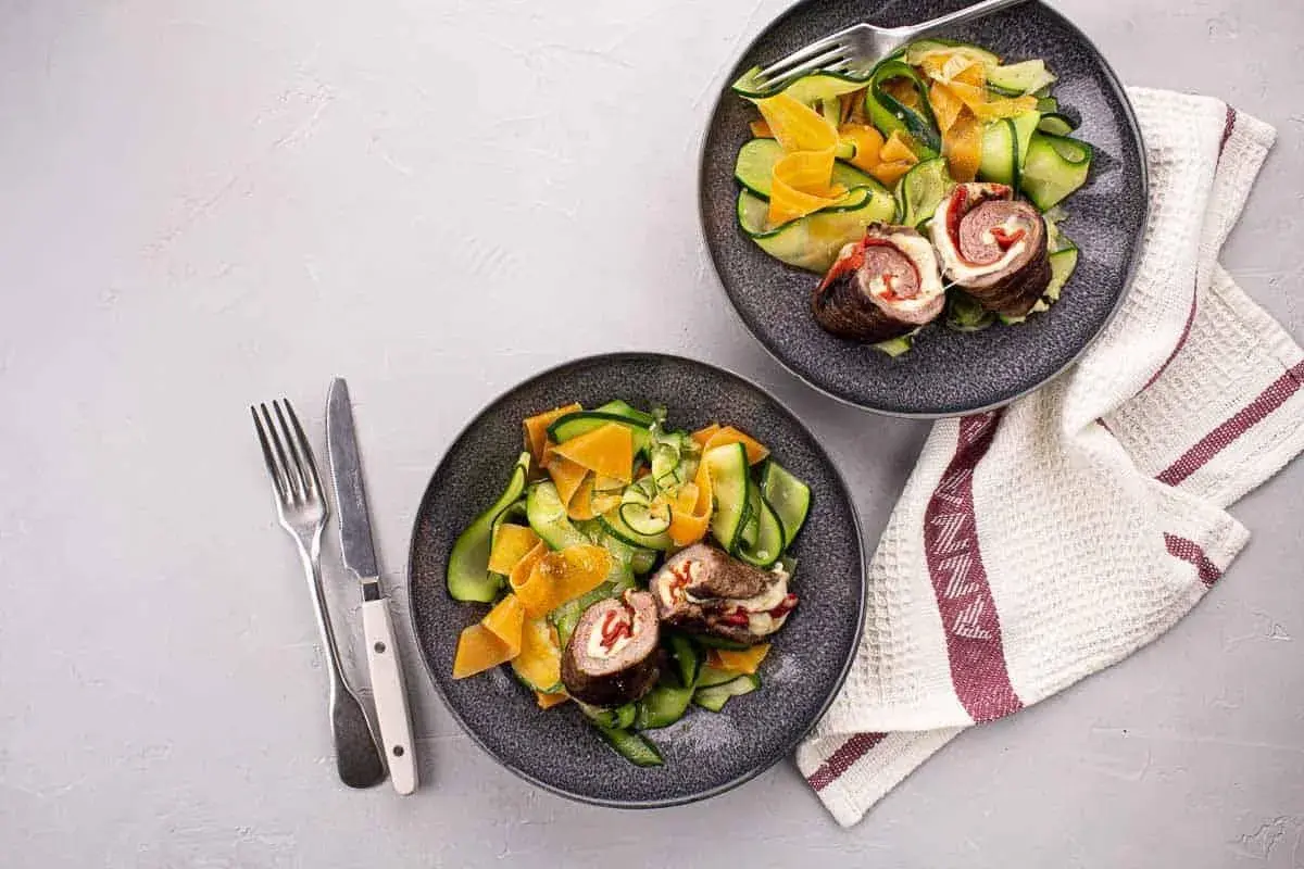 Deux assiettes de involtini, roulés de viande farcis, servis avec une salade de courgettes et carottes en rubans.
